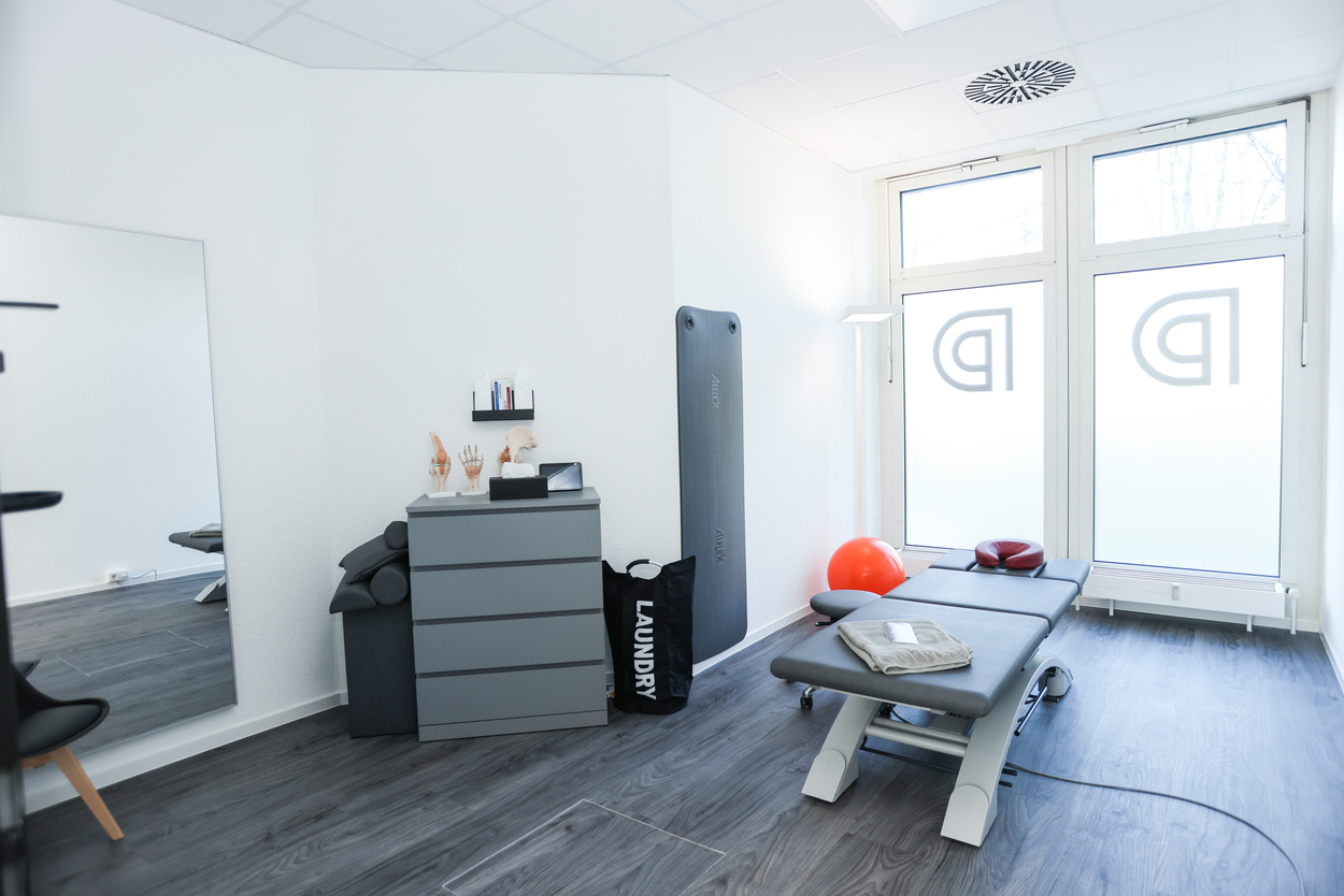Heller Physiotherapieraum mit Behandlungsliege, Spiegel, Schrank; helle Wände und Holzfußboden.