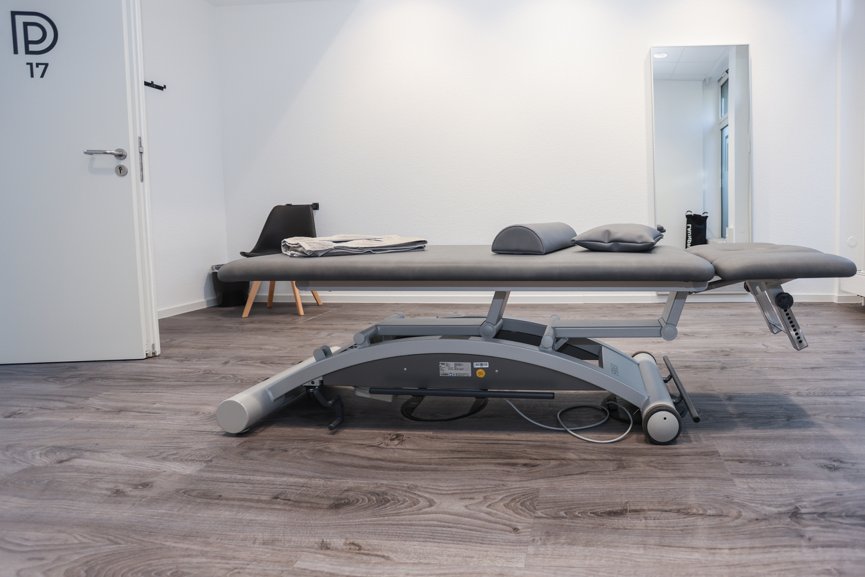 Behandlungszimmer mit grauer Liege, hellem Holzboden und modernen Dekorationselementen.