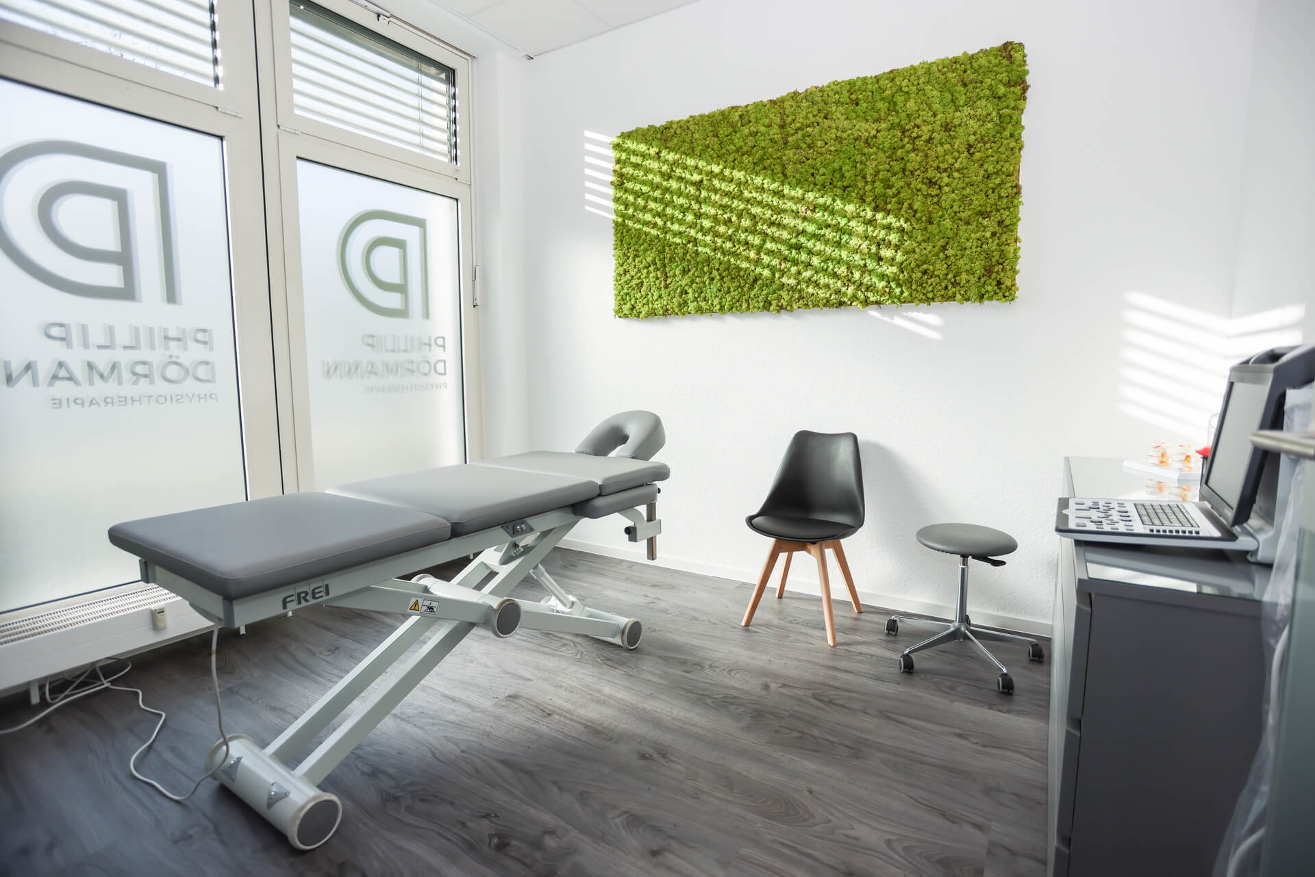 Modernes Behandlungszimmer mit grauem Behandlungstisch, grünem Moosbild und vier Stühlen.