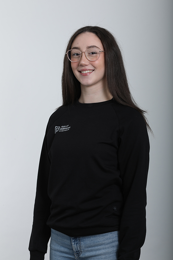 Frau mit Brille lächelt leicht, trägt einen schwarzen Pullover und blaue Jeans.