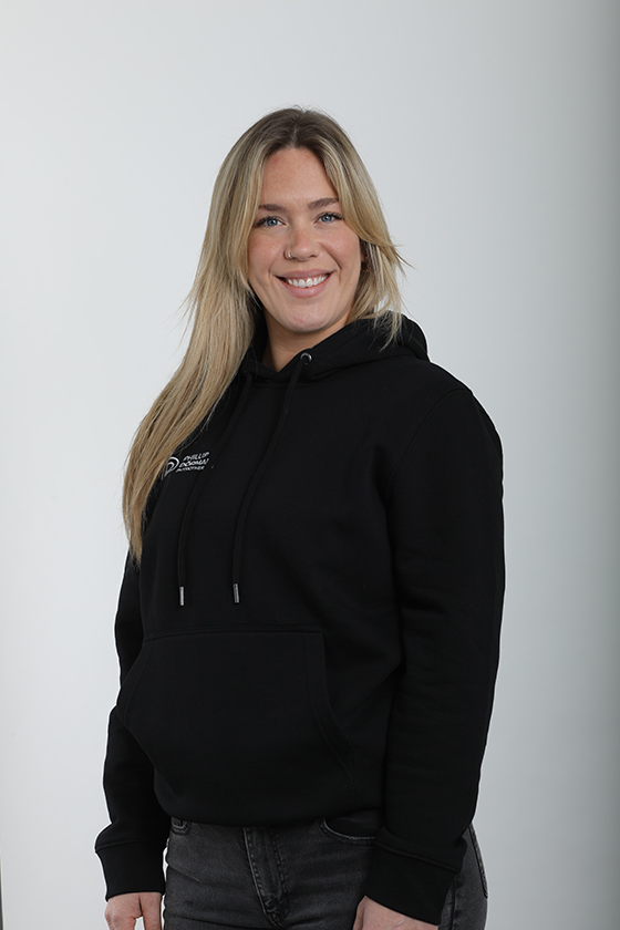 Eine lächelnde Frau mit blondem Haar, trägt einen schwarzen Hoodie vor weißem Hintergrund.
