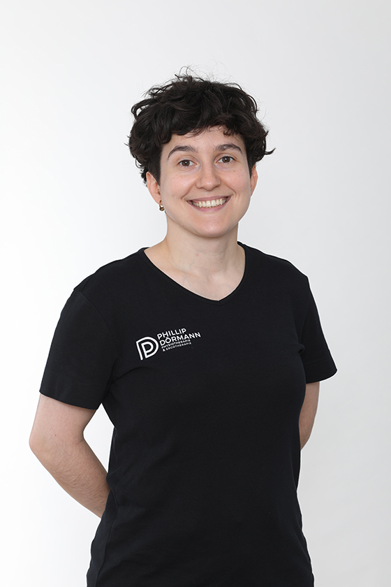 Person mit dunklen, lockigen Haaren, lächelt, schwarzes T-Shirt mit Logo drauf.