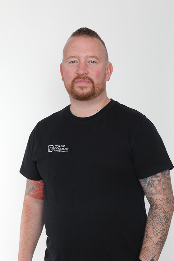Ein Mann mit Bart lächelt dezent, trägt ein schwarzes T-Shirt und zeigt tätowierte Arme.