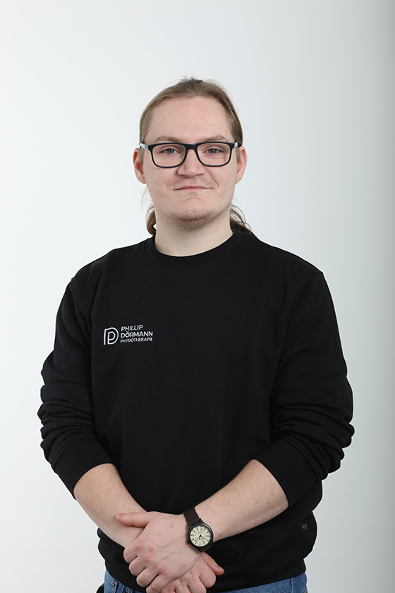 Eine Person mit Brille und schwarzem Pullover, lächelnd, mit Armbanduhr, vor weißem Hintergrund.