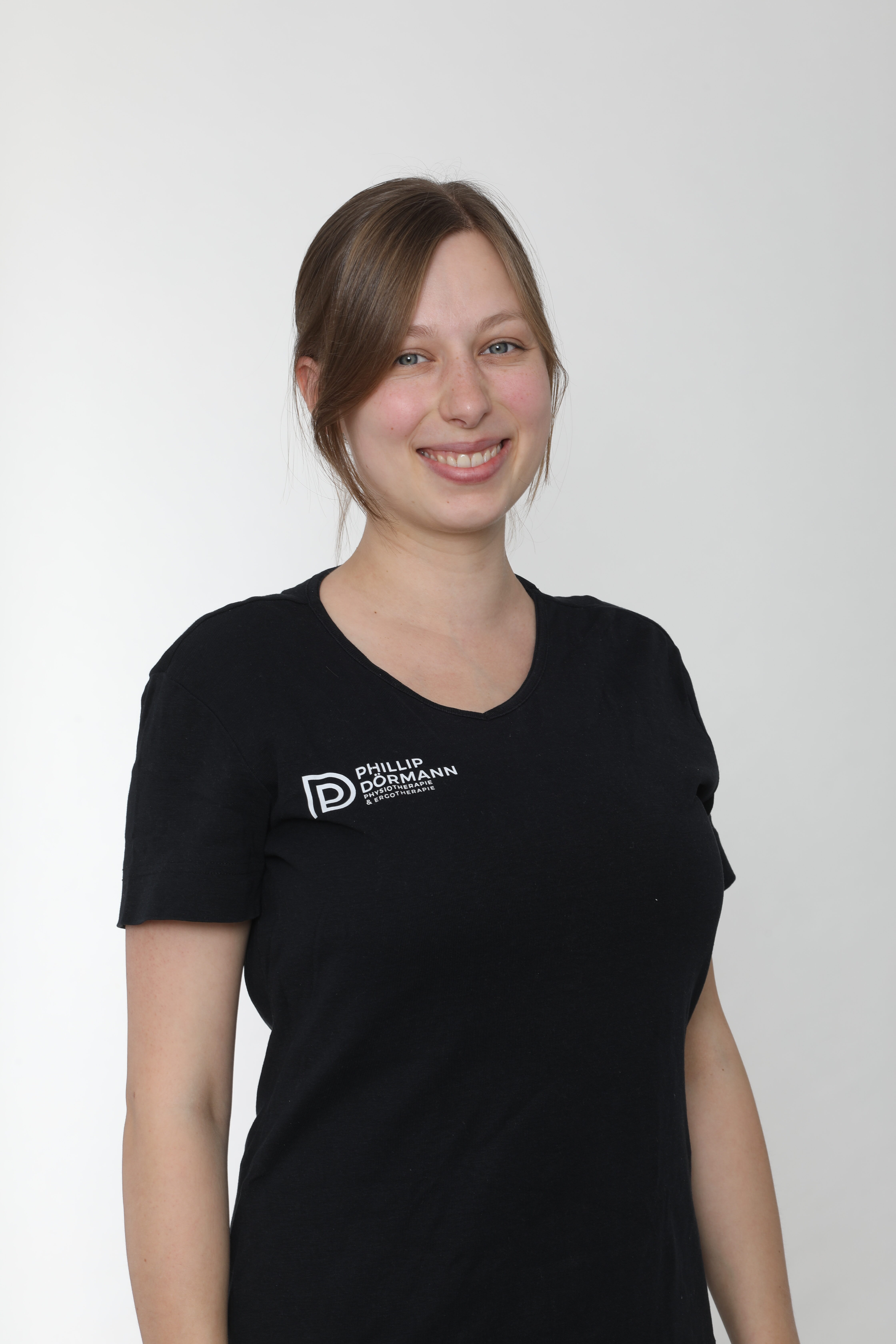 Frau mit braunem Haar lächelt, trägt schwarzes T-Shirt mit Logo auf weißem Hintergrund.