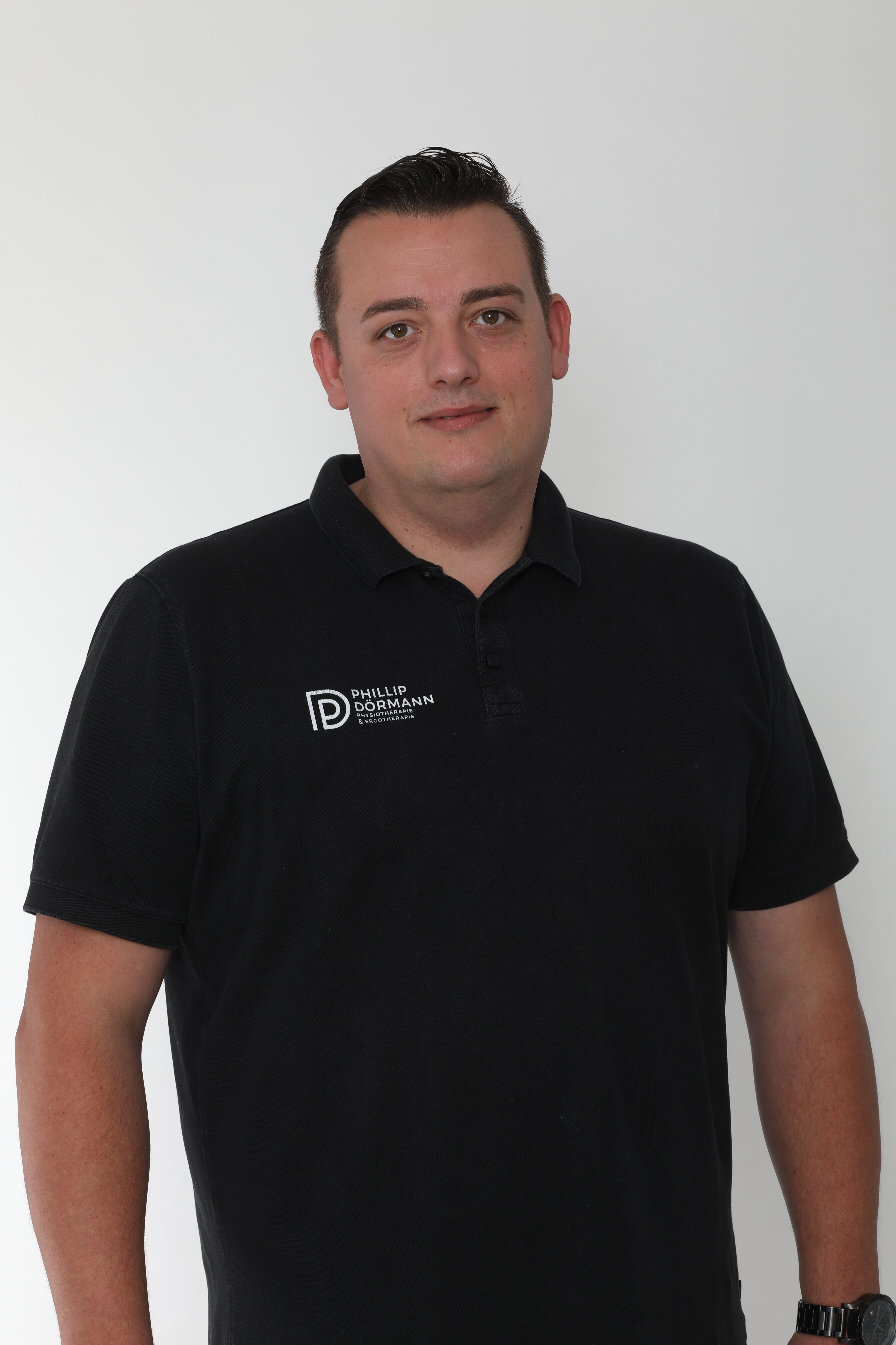 Mann in schwarzem Polohemd mit Firmenlogo, neutraler Ausdruck, heller Hintergrund.