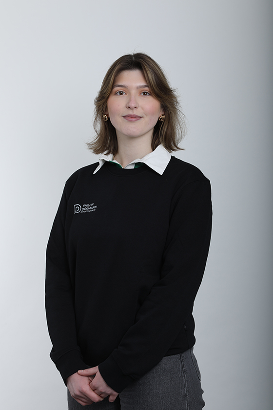 Eine lächelnde Frau in schwarzem Pullover mit Logo, vor grauem Hintergrund.