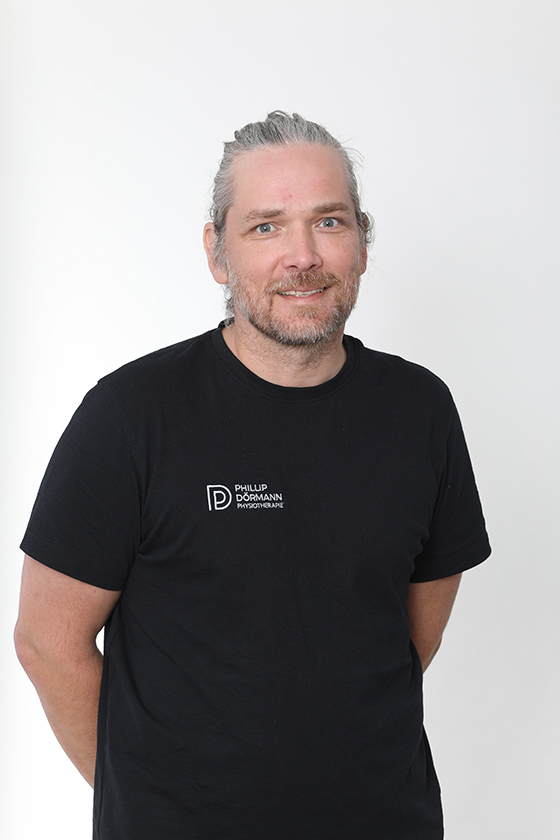 Graubärtiger Mann lächelt in schwarzem T-Shirt vor weißem Hintergrund.
