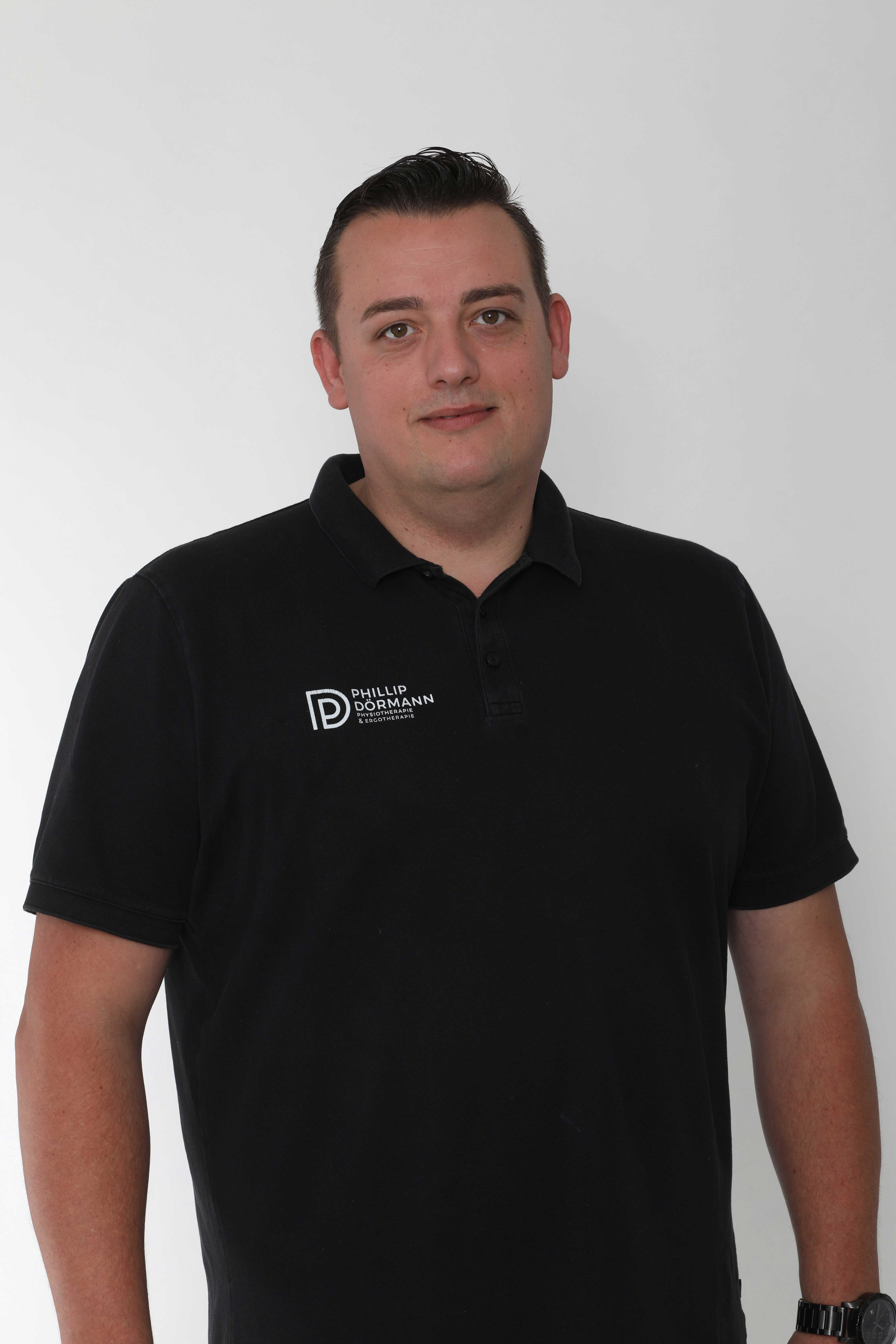 Mann in schwarzem Polohemd, neutraler Gesichtsausdruck, kurzer dunkler Haarstyle.