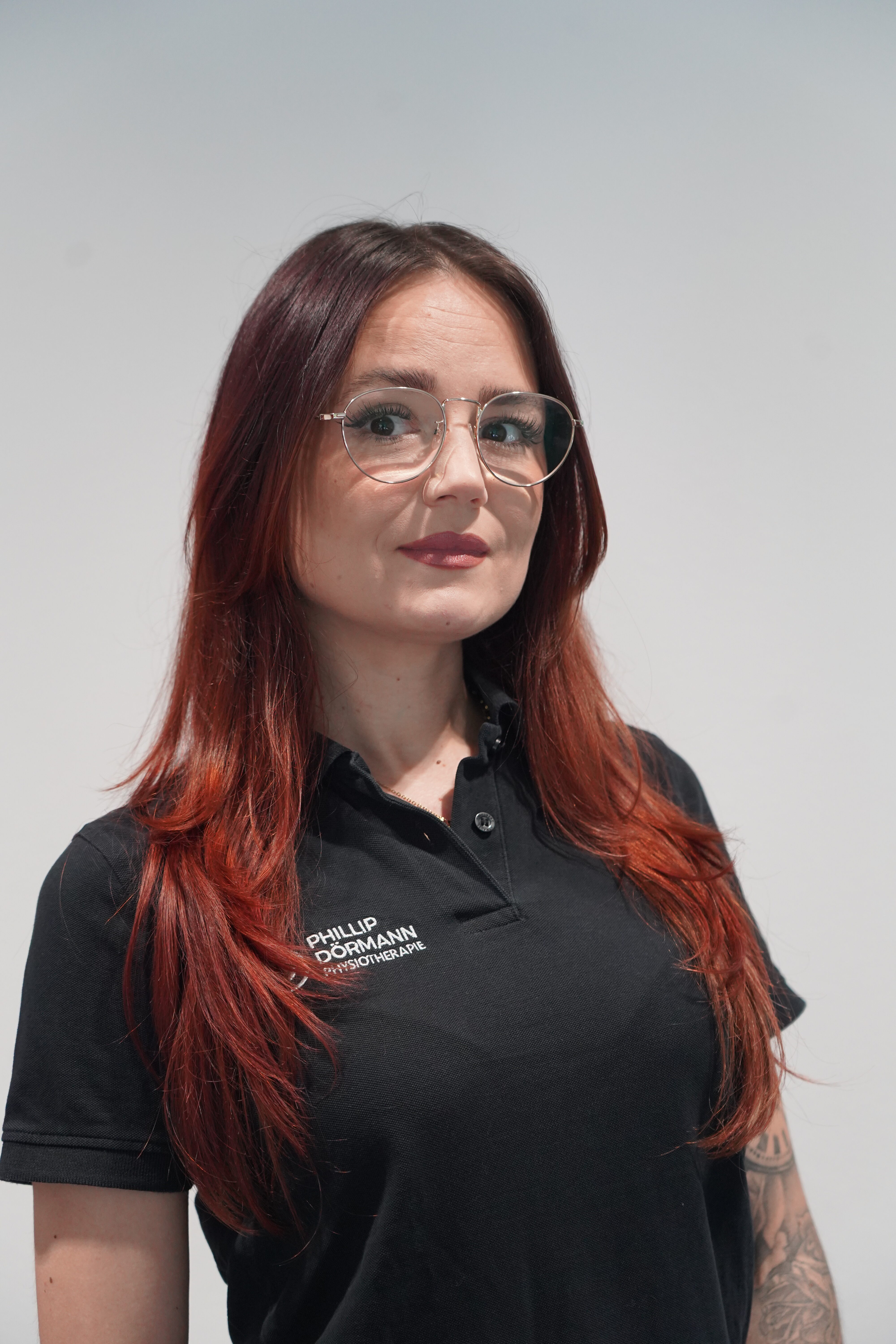 Frau mit neutralem Blick, rotem Haar, Brille und schwarzem Polo mit Logo