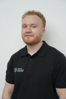 Junger Mann mit rotblonden Haaren und Bart in schwarzem Poloshirt laechelt